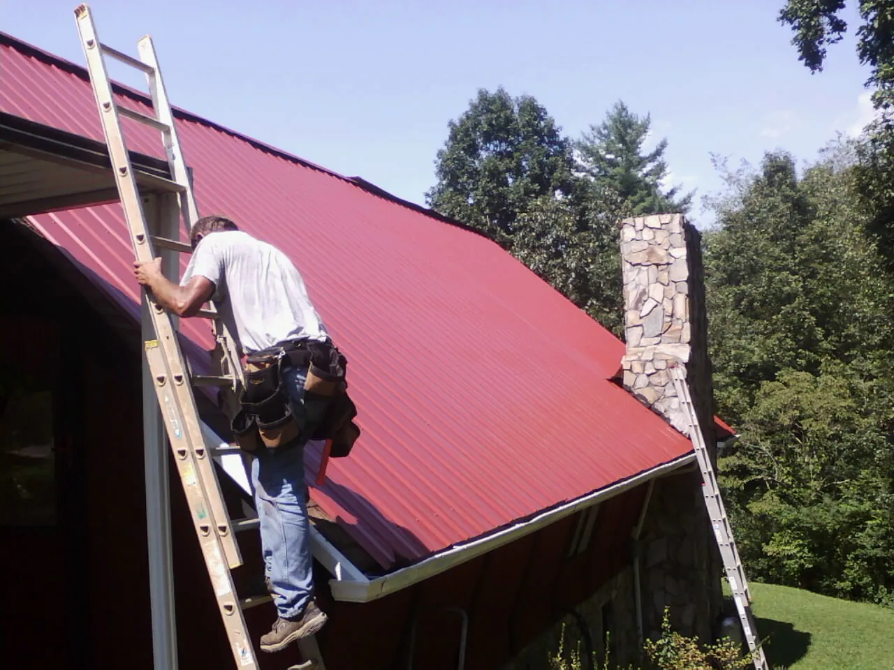 Roofer on a ladder beside a red metal roof and stone chimney — Soffit & Fascia Repair by Roofing Brothers in Craig